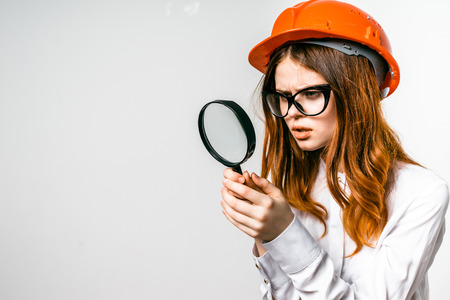 Young woman builder in big glasses in a hard hat looks into a magnifierの写真素材