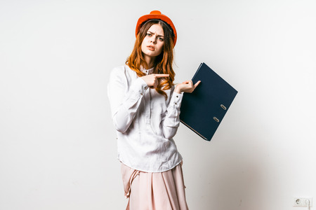 A young girl in an orange hat with doubt points to a folder with documents. Isolated on white backgroundの写真素材
