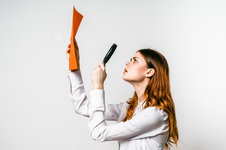red-haired girl in a white shirt looks at an orange sheet A4 through a magnifying glassの写真素材