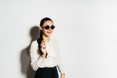 smiling girl in glasses and a white blouse holding a gold coin and looking to the side, crypto currency, bitcoins, isolated on a white backgroundの写真素材