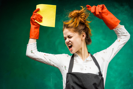 an aggressive red-haired girl in a large apron and wearing orange rubber gloves holds a yellow ragの写真素材