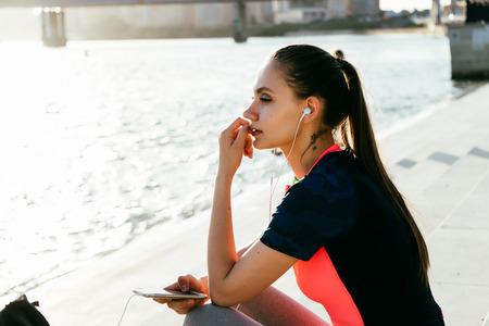 a girl in a tracksuit sits on the riverbank after a jog and listens to music on headphonesの写真素材