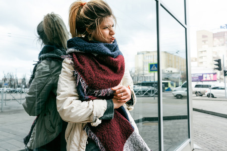 a girl with a big scarf stands on the street of the city, leaning against a mirror wall and looking...