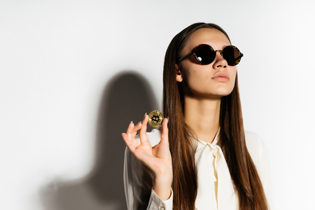 a young modern successful business lady in sunglasses holds a gold bitcoin in her hands, thinks about the crypto currencyの写真素材