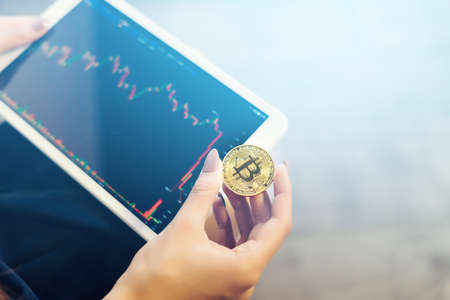 modern successful girl holds a gold bitcoin and a tablet with charts of crypto currencyの写真素材