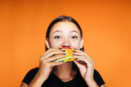 Happy girl on an orange background eating her burgerの写真素材