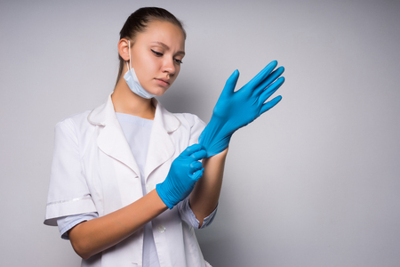A sad girl in a white coat and wearing a mask puts on gloves. Isolated on white backgroundの写真素材