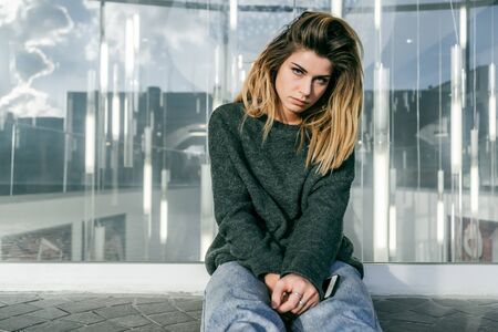 sad stylish girl with thick hair sitting near a glass wallの写真素材