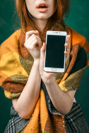 red-haired young girl shows the screen of her smartphone, checkered orange scarf on her shouldersの写真素材