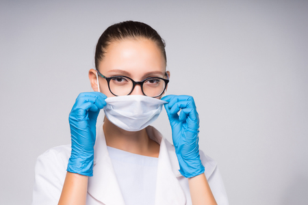 intelligent young woman doctor wearing glasses and white protective mask on face, wearing blue gloves looking at cameraの写真素材