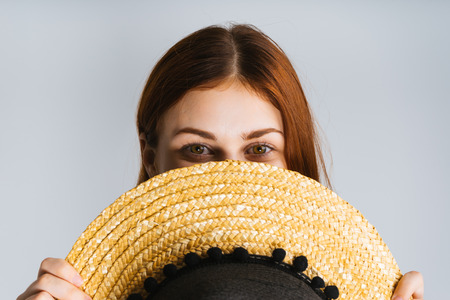 young red-haired girl hides her face behind a straw hat, over white background