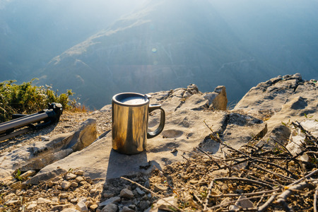A metal mug stands on the edge of the cliff, against the background of high mountains, in the rays of the sunの写真素材