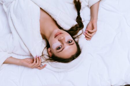 a young girl lies in bed in a white coat, just woke up, under the eyes of silicone white patches against bruisesの写真素材