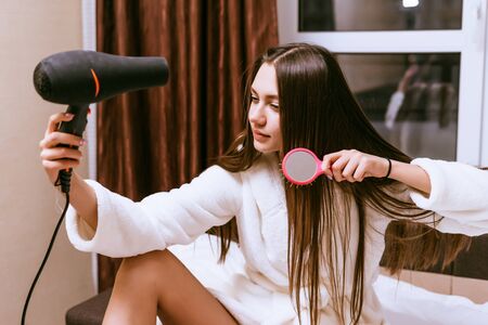 a young long-haired girl in a white terry robe dries her hair with a hairdryerの写真素材