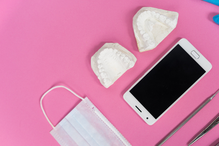 on the pink surface lie a white smartphone, a medical face mask, a plaster cast of teeth and dental...