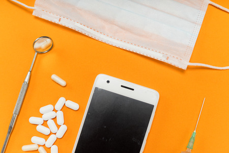 on an orange background lie a white smartphone, tablets, a protective medical mask, a syringe and dental instrumentsの写真素材