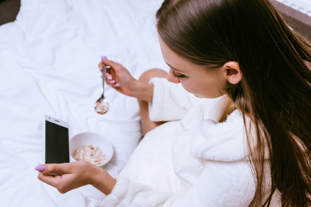 a young girl in a white terry dressing gown is having breakfast in bed, looking into her smartphone