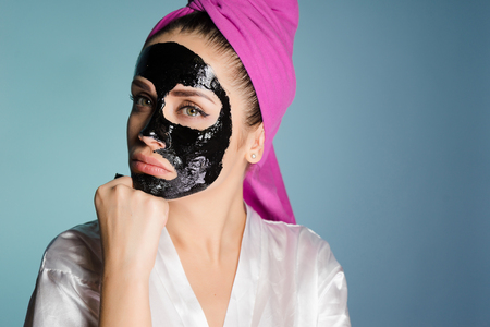 a young pensive girl with a pink towel on her head applies a black cleansing mask on her faceの写真素材