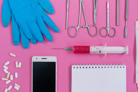 on a pink surface lie medical instruments, blue gloves, a smartphone, a notebook,white tablets