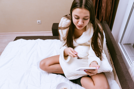 a cute young girl in a white terry dressing gown is sitting on the bed, having breakfast and looking into the phoneの写真素材