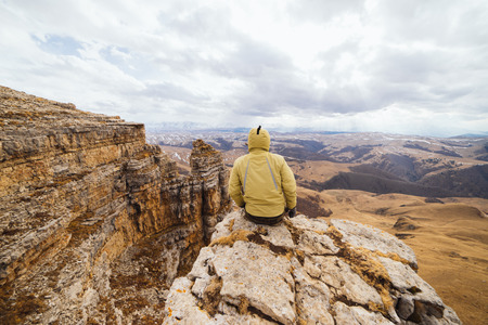 male traveler in a yellow jacket sits on the edge of a cliff and enjoys nature and mountainsの写真素材