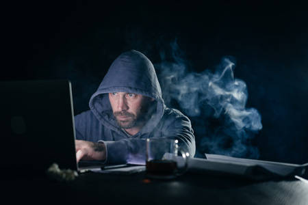 secretive mysterious man hacker in hood hides his face doing something illegal on laptopの写真素材
