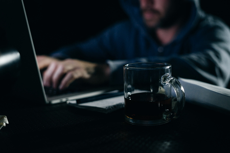mysterious criminal man in a hood hacking something on a laptop in the dark