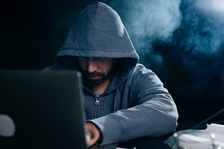 young guy hacker in a sweatshirt with a hood sits behind a laptop