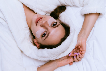 a surprised young girl lies on a bed, in a white terry dressing gown, under the eyes of a patchの写真素材