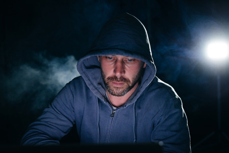Mysterious bearded man in a hood hacks a laptop in the dark
