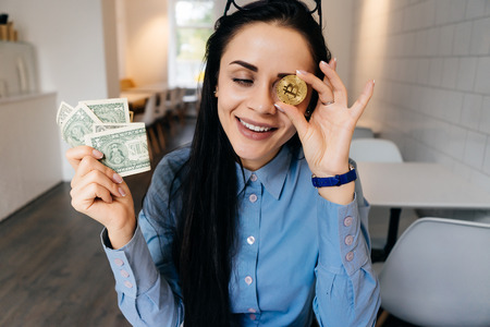 Laughing young girl in blue shirt is holding golden bitcoin and dollars in handsの写真素材