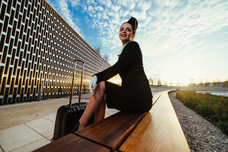 slender young woman stewardess sitting on bench in park, with suitcase, waiting for her flightの写真素材