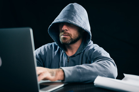 young guy hacker in a sweatshirt with a hood sits behind a laptopの写真素材