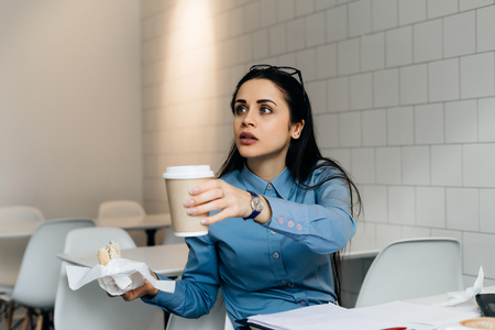 Young girl student in a blue shirt eating breakfast in a cafe, holding a sandwich and coffeeの写真素材