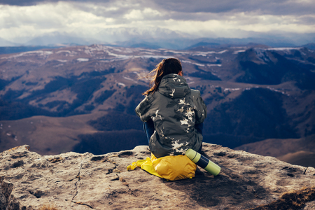 young girl with a yellow backpack sits on the edge of a cliff, enjoying the mountain natureの写真素材