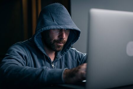 a man hacker sits in a room behind a laptop and prints somethingの写真素材