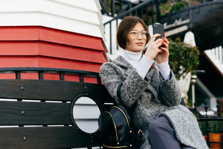 beautiful stylish girl in a gray coat and glasses sits on a bench in the street and takes picturesの写真素材
