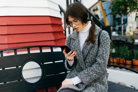 stylish young dark-haired girl with glasses and a gray coat writes a message on the smartphoneの写真素材