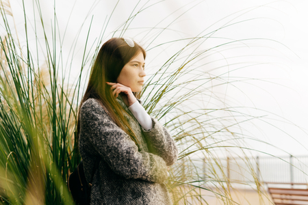 young dark-haired girl in gray coat walking through city streets, posing near green plantsの写真素材