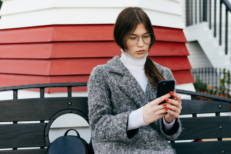 stylish young dark-haired girl in a gray coat and glasses sits on a bench outside and writes a message on the smartphoneの写真素材