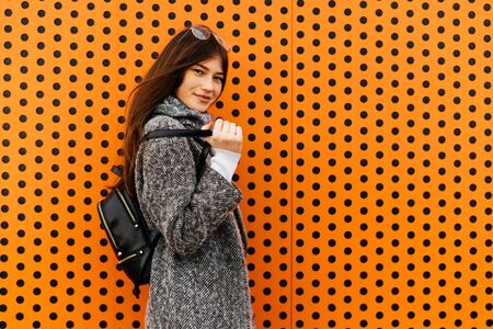 beautiful dark-haired girl in gray coat walking on city streets, posing against an unusual orange...