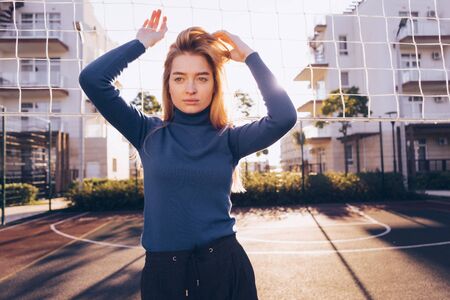 stylish beautiful blond girl in a blue sweater posing on a football field outdoors in the sun