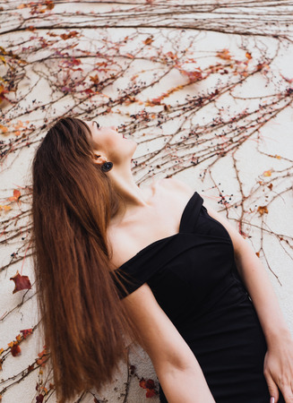 luxurious long-haired young woman in a black dress is standing by the white wall and looking upの写真素材