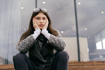 sad young brunette girl in gray coat sits on outdoor chairs