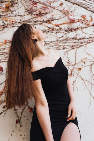 luxurious long-haired woman in a black dress posing against an unusual wall background, looking upの写真素材