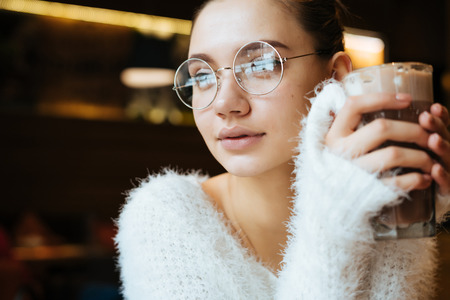 cute young girl student wearing glasses and a white jacket sitting in a cafe and drinking a delicious latteの写真素材