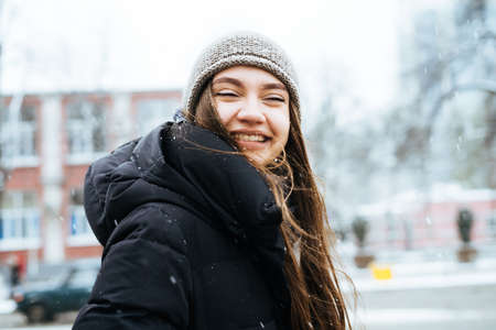 happy young long-haired girl in a hat and warm jacket walking through the streets of the city and smilingの写真素材