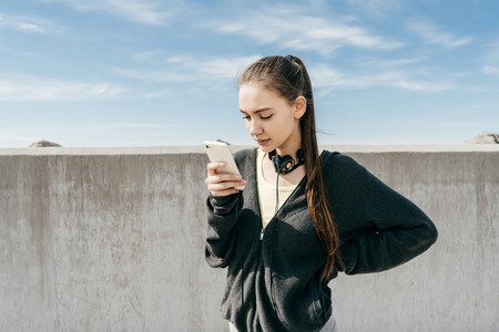 stylish young girl stands outdoors under a blue sky, looks into the phoneの写真素材