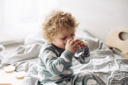 cute little baby, boy, with curly hair sitting on the bed and drinking waterの写真素材