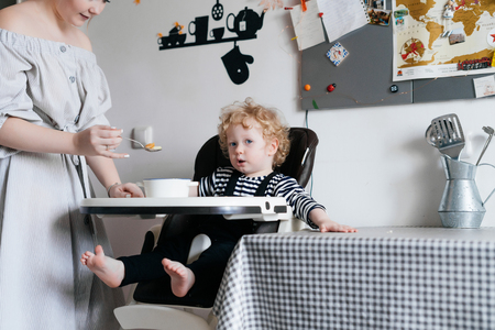 family is having breakfast, a caring young mother is feeding her little baby, the boy is sitting in a child's chair in the kitchenの写真素材